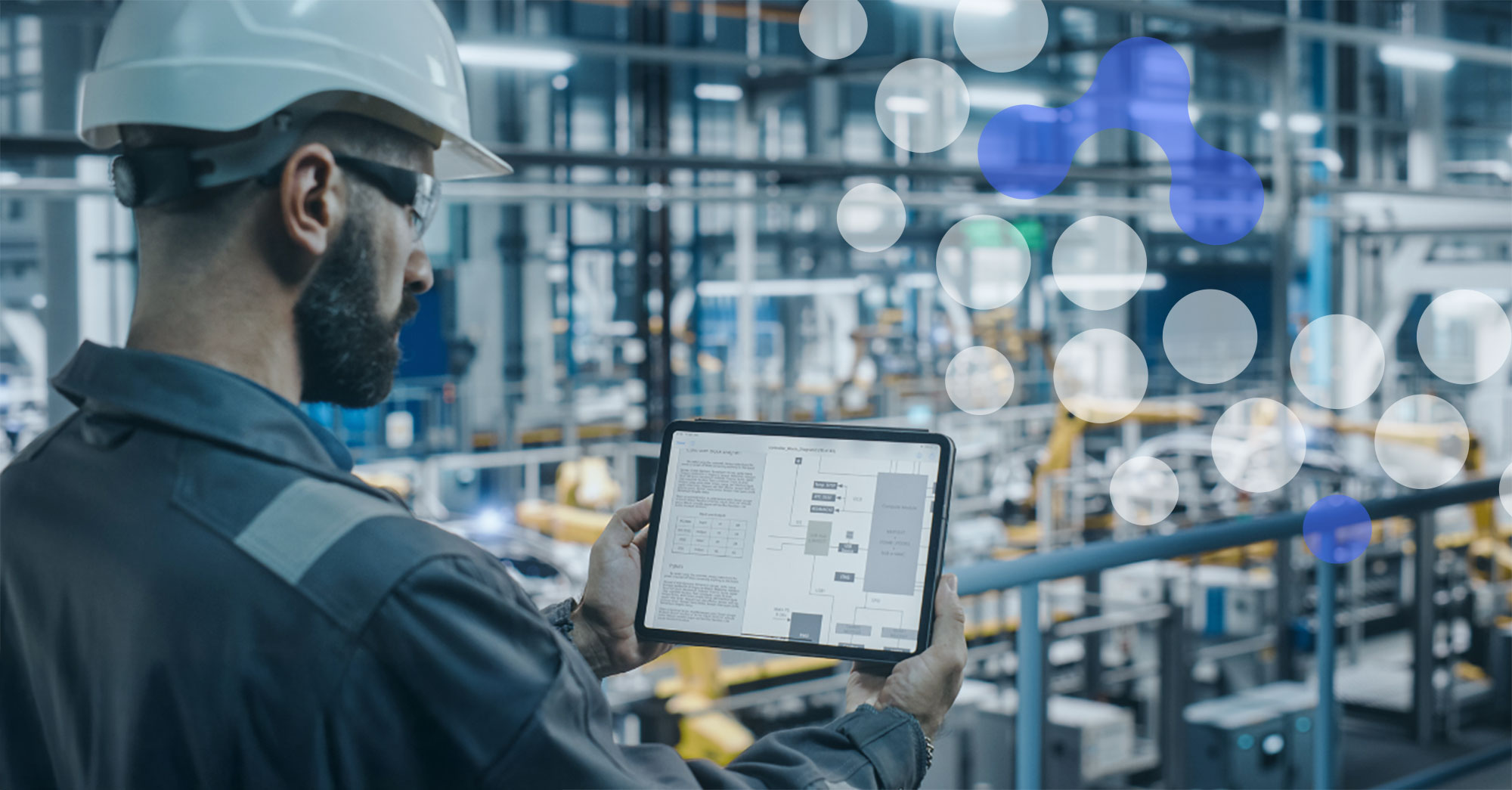 A worker in a hard hat and eye protection, surrounded by pipes, holds up a tablet to review a schematic on the screen.